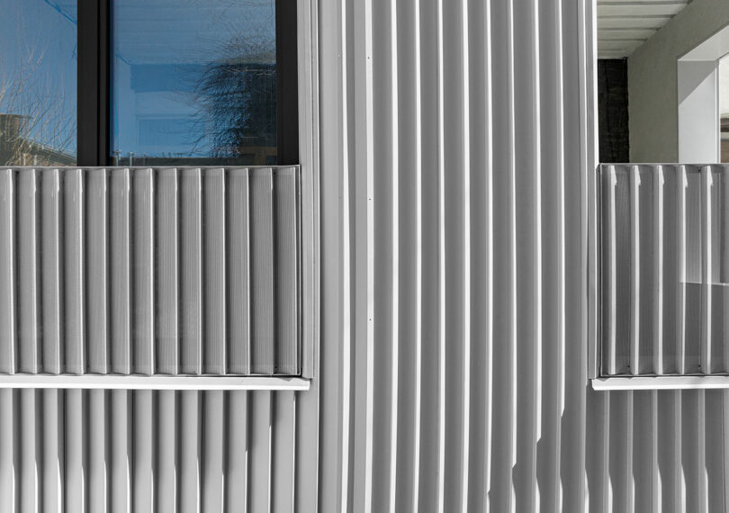 Close-up of a modern building facade featuring vertical metallic ridges and large glass windows, reflecting part of an outdoor scene.