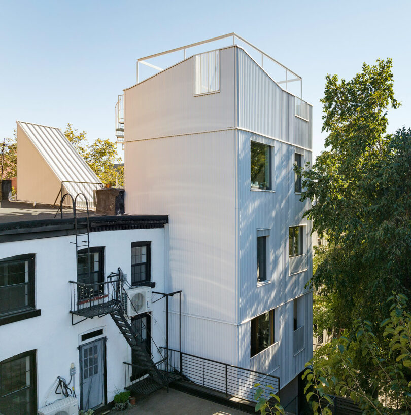 A modern three-story white building with large windows stands next to an older white building with a black fire escape staircase and metal railing. Trees and a clear sky are visible in the background.