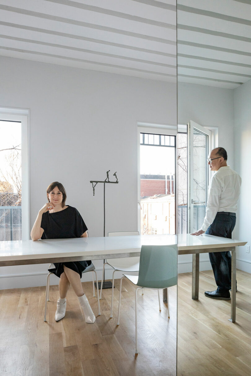 A woman sits at a table in a bright room with white walls, while a man stands near an open door. An outdoor view can be seen through the large windows and open door.