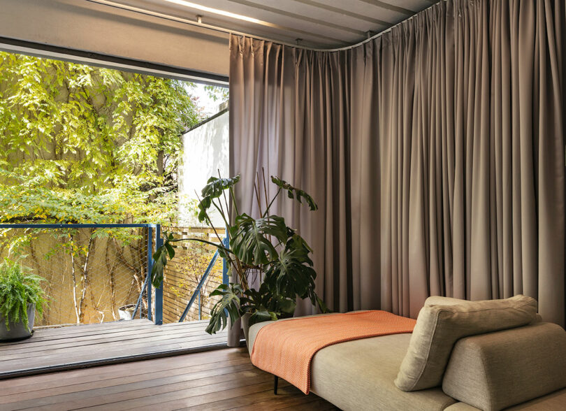 A serene indoor space with a beige daybed, grey curtains, and a view of lush greenery outside a glass door. A potted plant sits next to the door.