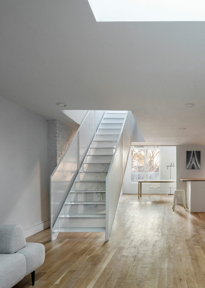 A modern, minimalist interior featuring a white staircase with glass railings, light wooden flooring, a small desk by a window, and a skylight above.