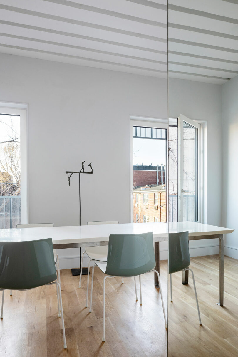 A minimalist room with a long white table, three modern chairs, wooden floor, two windows, and a door leading to a balcony. A sculpture of two figures perched on a branch is visible on the table.