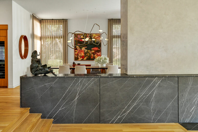 Modern interior with a marble counter, abstract chandelier, wall art, and a contemporary sculpture exuding Brazilian Modernism. Light-colored drapes cover large windows, and a dining table with chairs is visible in the background.