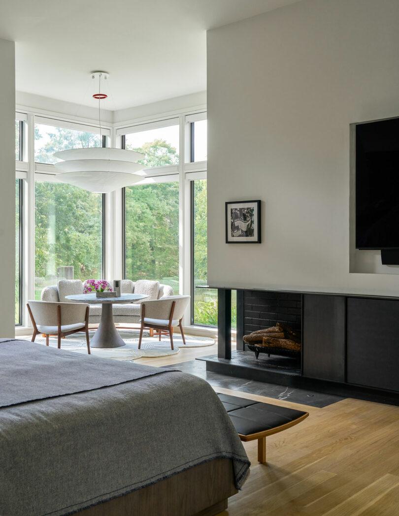 A modern bedroom showcasing Brazilian Modernism with a fireplace, large windows overlooking greenery, a seating area with a round table and four chairs, a wall-mounted TV, and a bed with a grey blanket on a wooden floor.