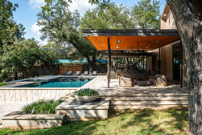 A modern outdoor patio with wooden flooring and a shaded seating area exemplifies Brazilian Modernism, set adjacent to a pool surrounded by greenery and lounge chairs under a clear sky.