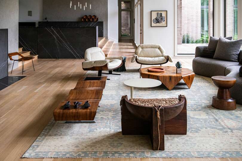 A modern living room with eclectic furniture including wooden coffee tables, armchairs, and a patterned rug that hints at Brazilian Modernism. The room features a large window and a living area with a black marble island in the background.