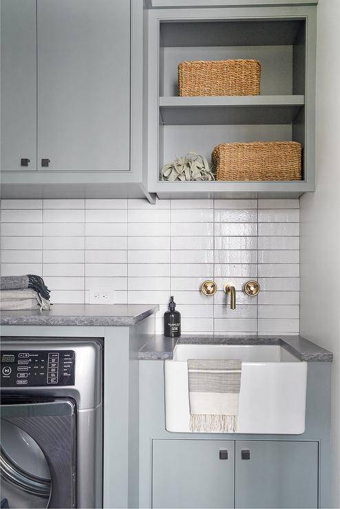 small white apron sink laundry room gold wall mounted faucet tile backsplash light gray cabinetry seagrass baskets