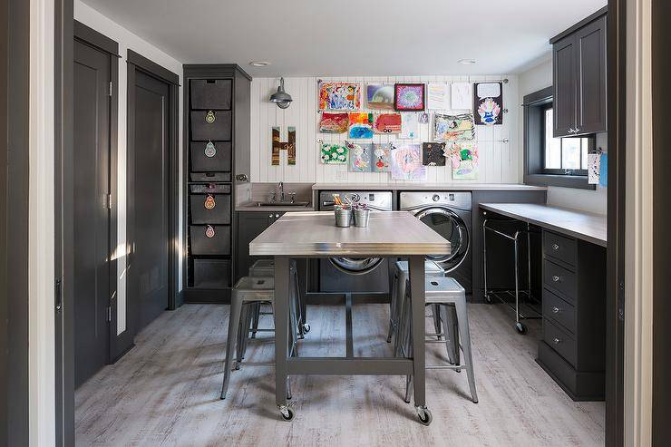 Tolix stools and a stainless steel rolling craft island centered in a gray laundry room doubling as a craft room. Black shaker cabinets frame the perimeter walls finished with white vertical planks displaying kids art on wires. Gray wash wood plank floors lighten the atmosphere of the room and creates a well balanced, decorative and practical space.