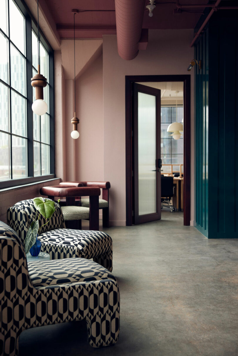 A modern interior featuring Malin patterned armchairs, a small table, and pendant lights. An open door leads to a conference room with another pendant light in view. Large windows line the left wall, flooding the space with natural light.