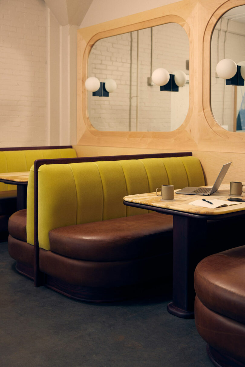 A cozy Malin coffee shop booth with green upholstery, a wooden table adorned with a laptop and two mugs, and a large mirror on the wall reflecting warm lights.