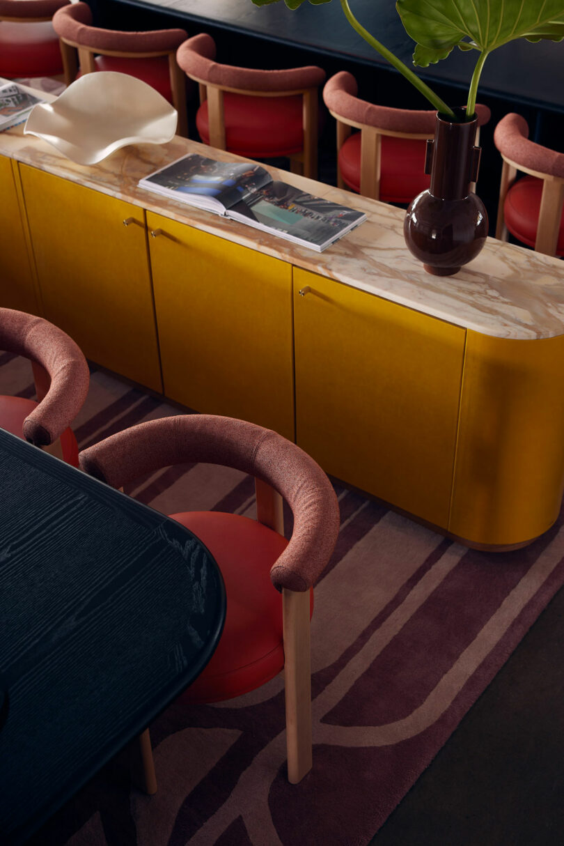 A modern dining area with a yellow marble-top sideboard, red and pink cushioned chairs, and a dark wooden table. There are open magazines and a brown vase with green leaves on the Malin sideboard.