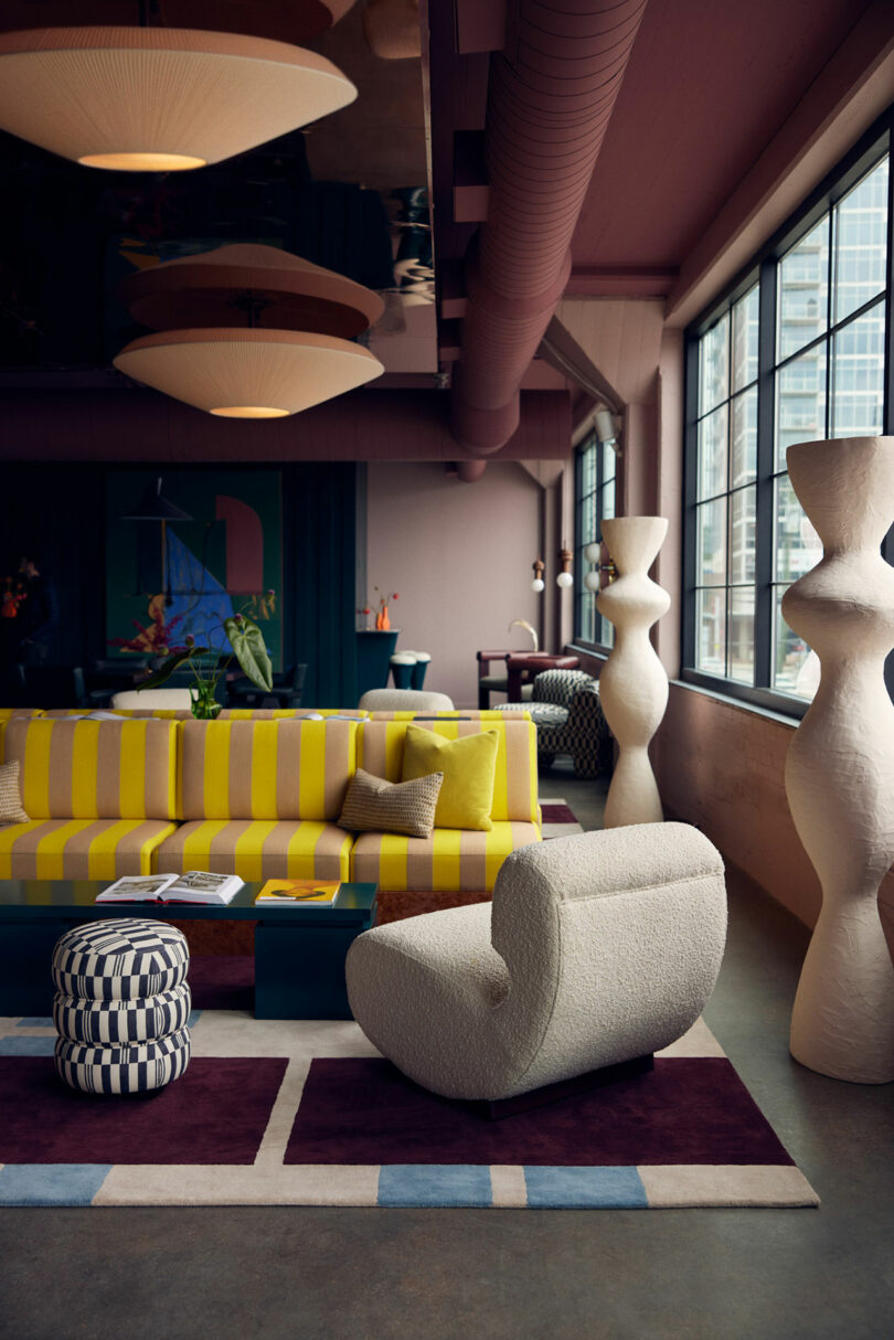 A modern living room features a yellow-striped sofa, a white Malin armchair, and geometric decor. Large windows allow natural light while colorful accents and unique sculptures add character.