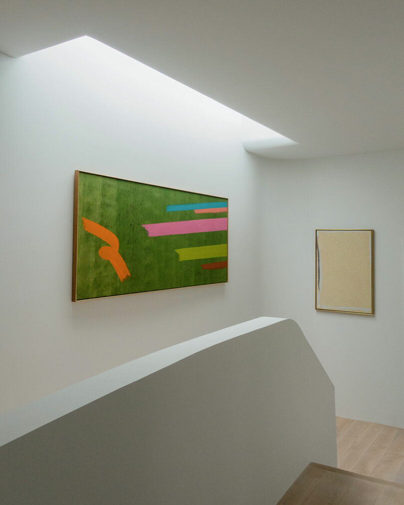 A minimalist indoor staircase with white walls, featuring a large abstract painting with vibrant colors on a green background on the left wall, and a simpler, neutral-toned painting on the right wall.