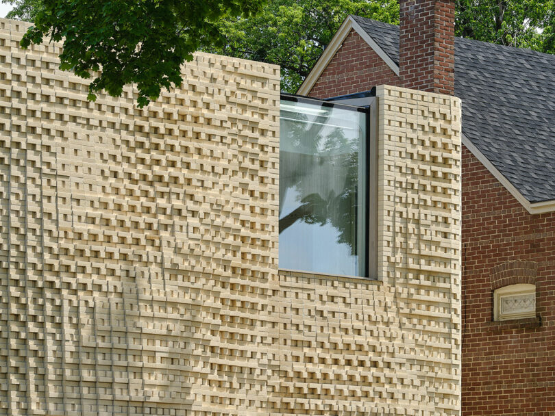 A modern brick extension with an irregular pattern adjoins a traditional brick house. The extension features a large window, and both structures are surrounded by greenery.