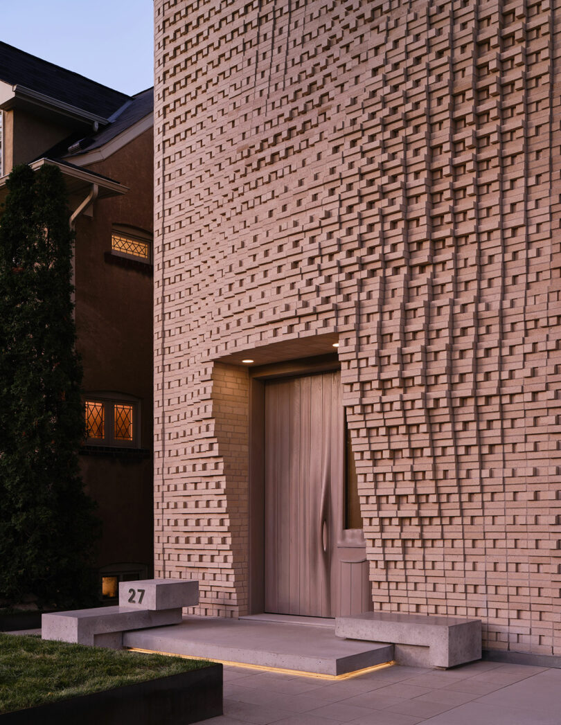 Modern brick building facade with a textured pattern, recessed wooden door, concrete steps and a bench labeled with the number 27. Adjacent building partially visible.