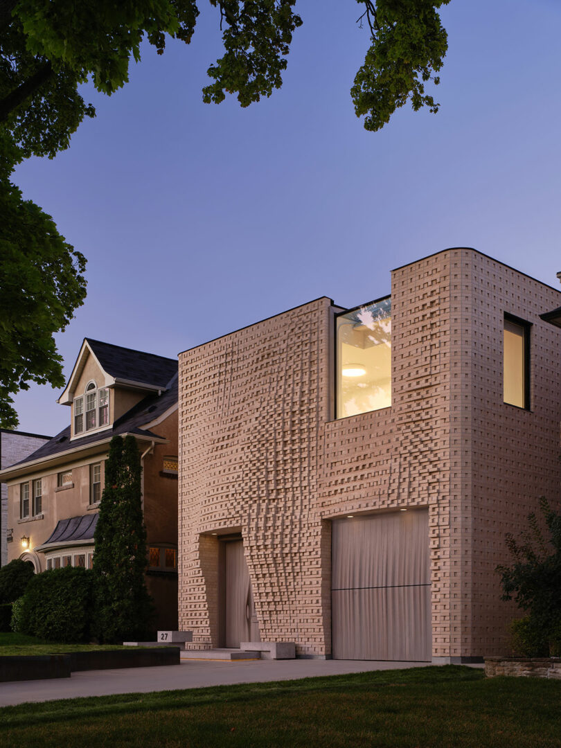 A modern two-story house with a unique textured facade and large window, adjacent to a traditional brick home, surrounded by trees.