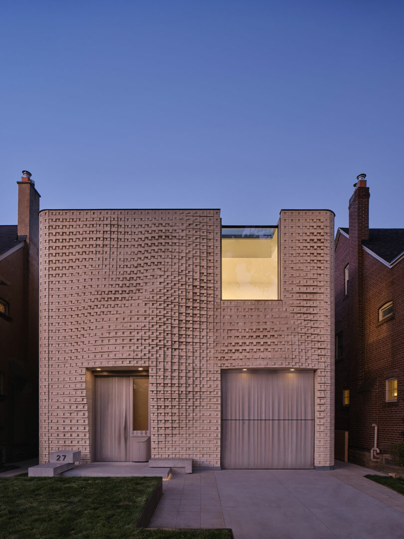 Modern two-story brick house with a unique textured facade, featuring a large illuminated window on the upper floor and a garage on the right. The house is flanked by two other buildings.