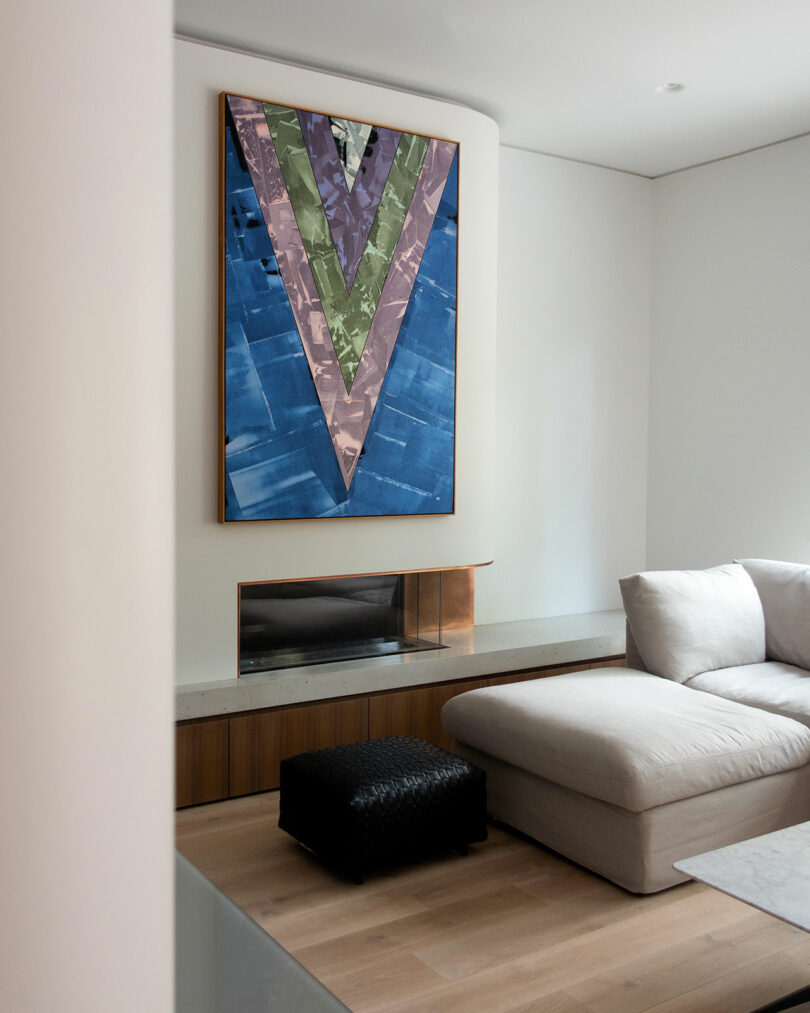 A modern living room with a minimalist design, featuring a large geometric artwork above a built-in fireplace, a cushioned sectional sofa to the right, and a black pouf in the center.