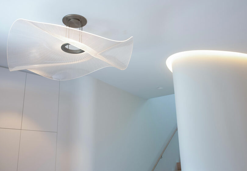 A modern, white, sculptural ceiling light fixture hangs in a minimalist room with white walls and a curved, softly illuminated column.