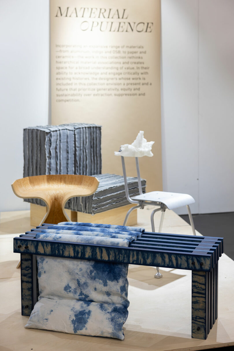 Exhibit showcasing modern furniture: a striped armchair, a wooden chair, and a metal chair with a white object on it. A blue-striped bench with two cushions serves as the launch pad for relaxation. Wall text titled "Material Opulence" in the background.