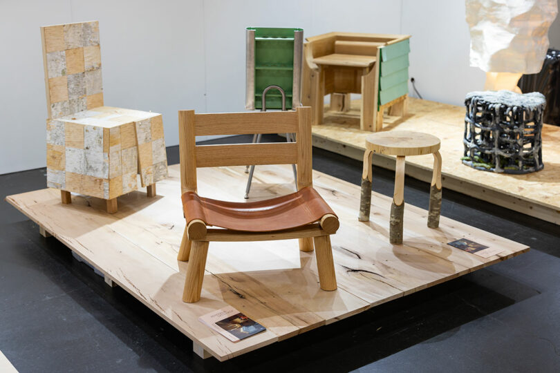 A display of wooden furniture pieces, including chairs, a stool, and a table, showcased on a wooden platform in a gallery setting serves as the perfect launch pad for artisanal craftsmanship.