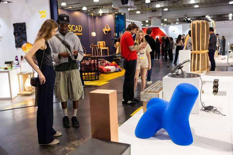 People view and discuss various modern art pieces displayed at an indoor exhibition. The space is well-lit with several visitors observing the exhibits, including a large blue abstract sculpture.