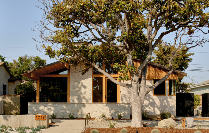 A modern, single-story house with wooden accents and large front windows, shaded by a large tree. The house number 7712 is visible on the mailbox.