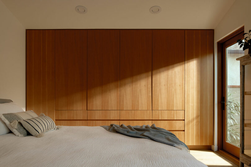 A bedroom with a neatly made bed featuring grey pillows and a blanket, a wooden wardrobe with vertical panels, and a sunlit patio door on the right.