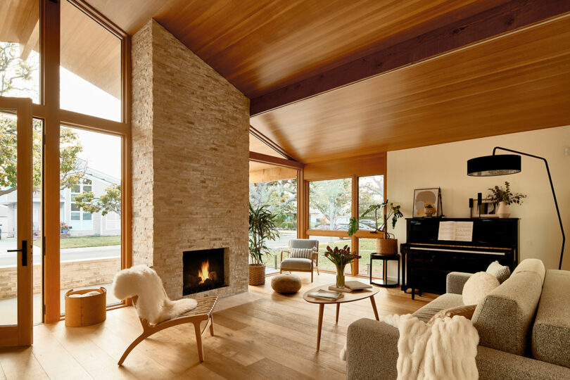 A cozy living room with a high wooden ceiling, large windows, a stone fireplace, modern furniture, a piano, and indoor plants.