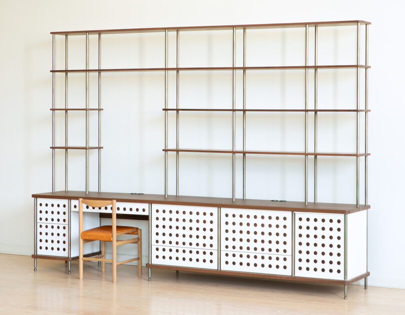 A modern shelving unit with multiple open shelves and a lower section of cabinets with perforated white doors, accompanied by a wooden chair with an orange cushioned seat.