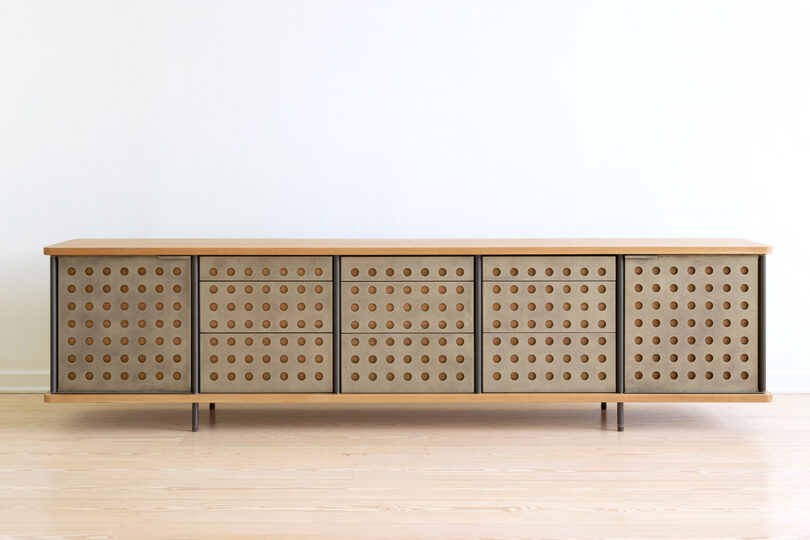 A long, modern sideboard with a light wood finish and perforated doors, standing on slim metal legs.