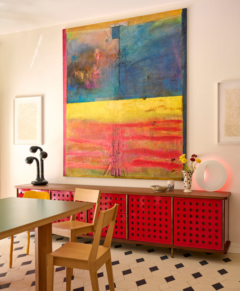 A modern dining room features a large abstract painting on the wall, a red perforated sideboard, a wooden table and chair, and a black sculptural piece. Geometric tile flooring complements the space.