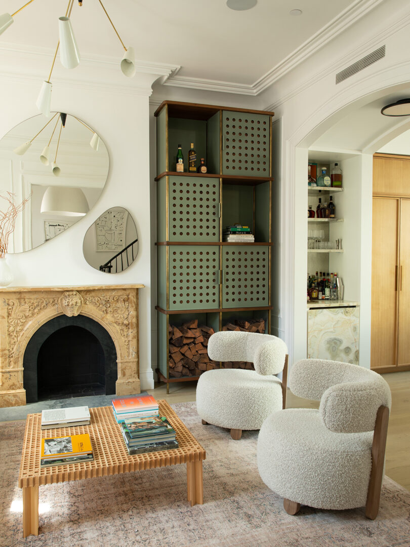 A modern living room features a fireplace, circular mirror, and a tall shelving unit. Two white chairs and a coffee table stand on a light rug.