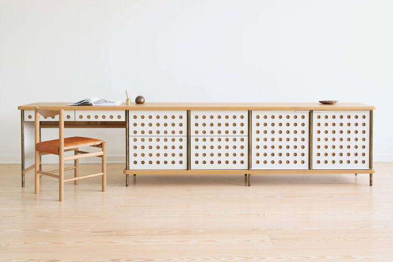 A minimalist wooden desk and chair with an orange cushion sit next to a perforated wooden cabinet against a plain white wall on a light wood floor.