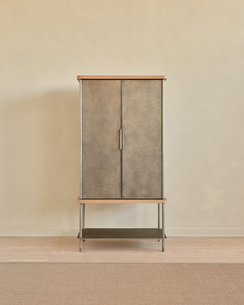 A tall, rectangular bar cabinet with two closed doors, a wooden top, and a lower shelf, positioned against a beige wall and standing on a light-colored floor.