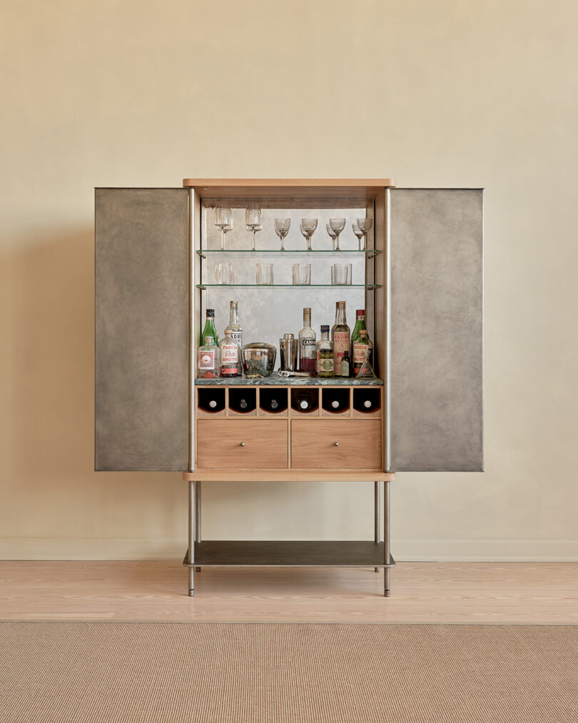 A tall, rectangular bar cabinet with two opened doors, a wooden top, and a lower shelf, positioned against a beige wall and standing on a light-colored floor.