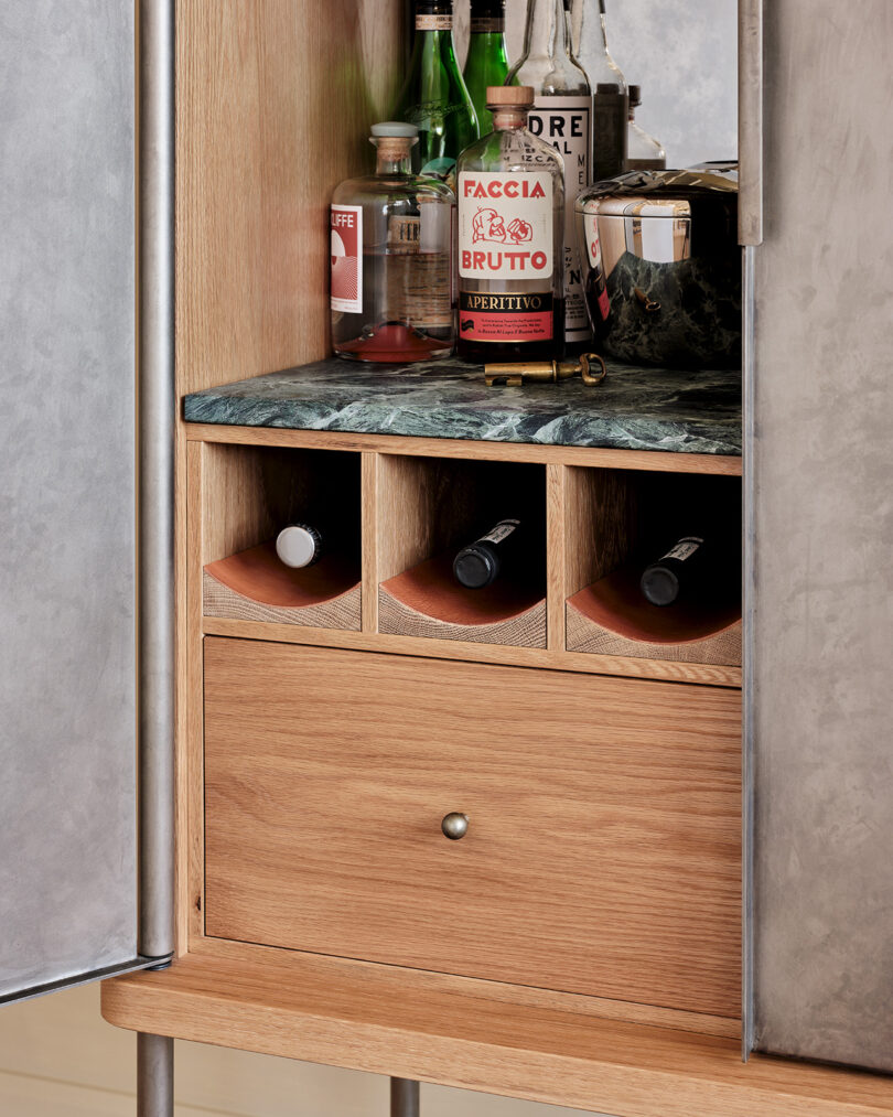 A wooden cabinet with an open door reveals bottles of alcohol, a cocktail shaker, and wine bottles stored horizontally in cubbies above a drawer.