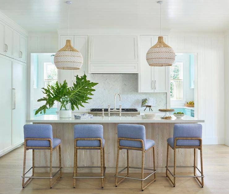 Blue and brown stools are placed at a cottage style island boasting a farmhouse sink and a polished nickel bridge style faucet lit by wicker pendant lights.