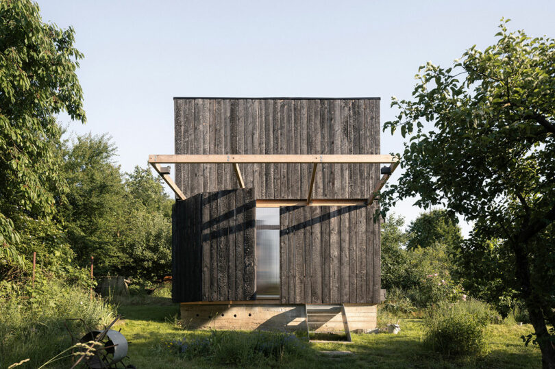 A small, modern cabin with dark wooden paneling and a reflective door, situated in a grassy area with trees.