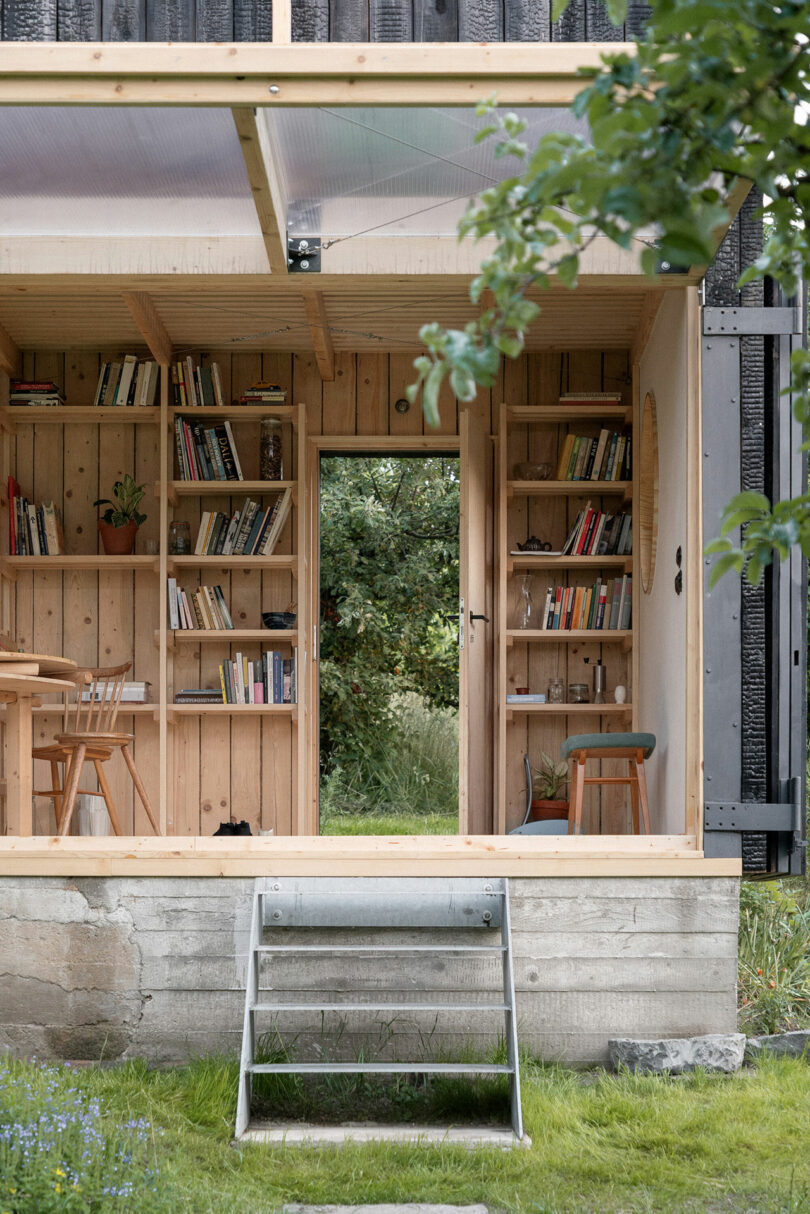 A small wooden cabin with open front wall revealing bookshelves, a table, and a stool. The cabin is surrounded by greenery with a metal staircase leading up to the entrance.