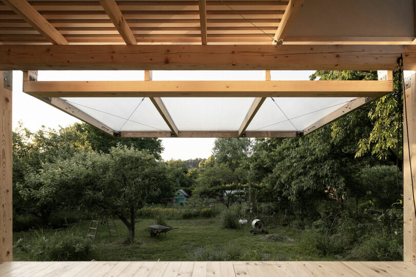 View of a lush garden with grass, trees, and a wheelbarrow, framed by the wooden structure and awning of an open building.