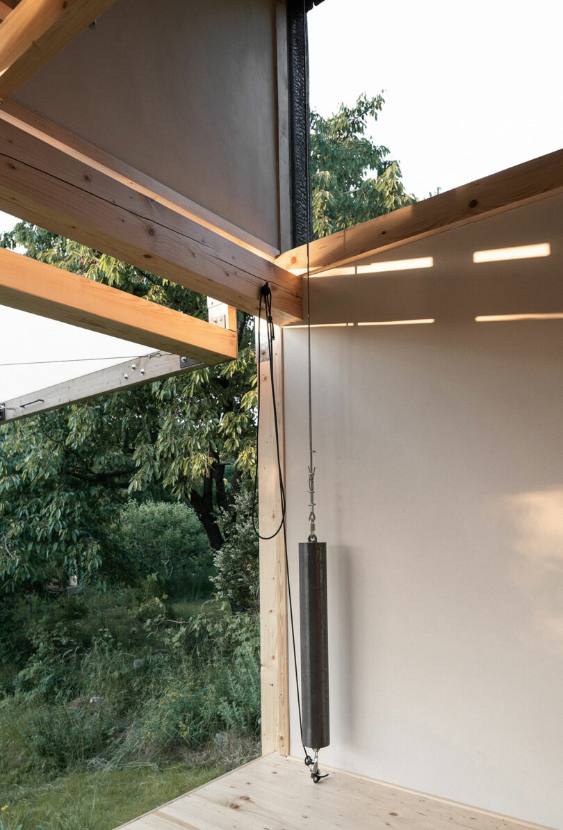 A wooden corner structure with a pulley system is attached to a cylindrical metal weight. Sunlight streams in, casting shadows on the adjacent white wall and revealing greenery outside.