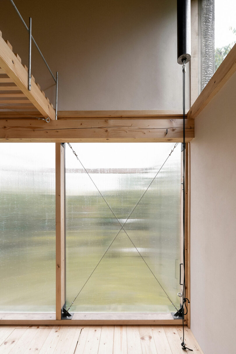 Interior view of a minimalist room with wooden floor, translucent corrugated panel walls, and a loft area accessed by a ladder. An open window offers a blurred view of greenery outside.