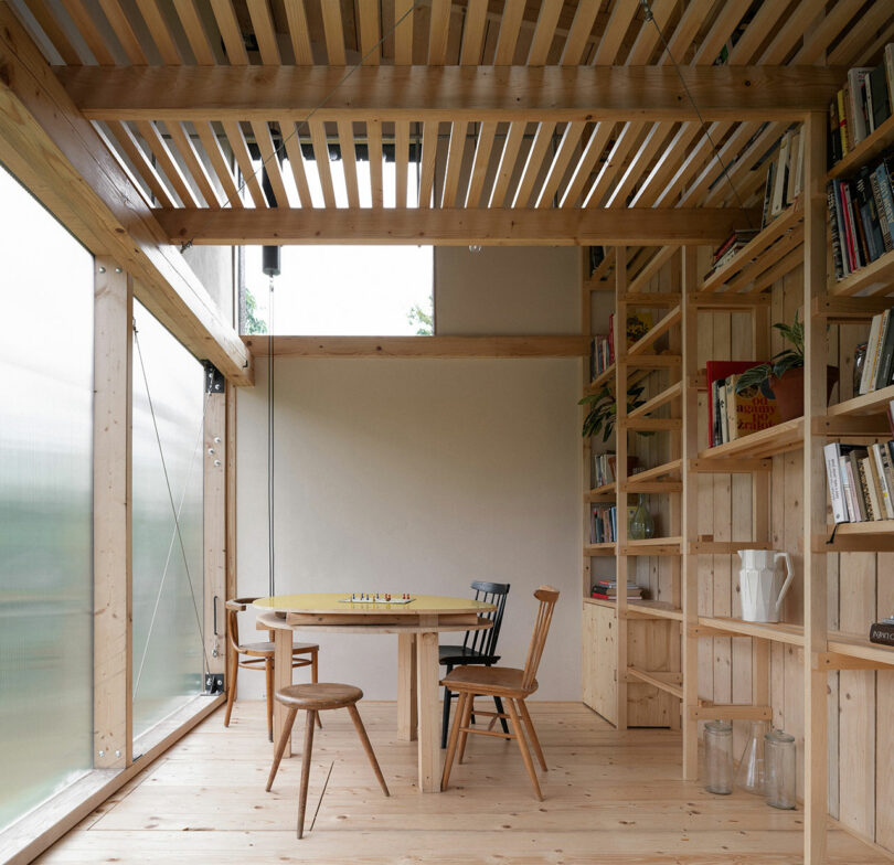 A wooden room with a round table and four chairs, surrounded by bookshelves and a large window bringing in natural light.