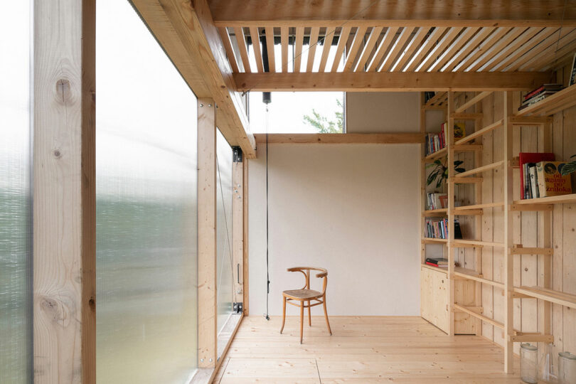 A minimalist room with wooden walls, floor, and ceiling. A bookshelf filled with books is on the right. A single wooden chair stands in the center. Large frosted windows allow natural light in.