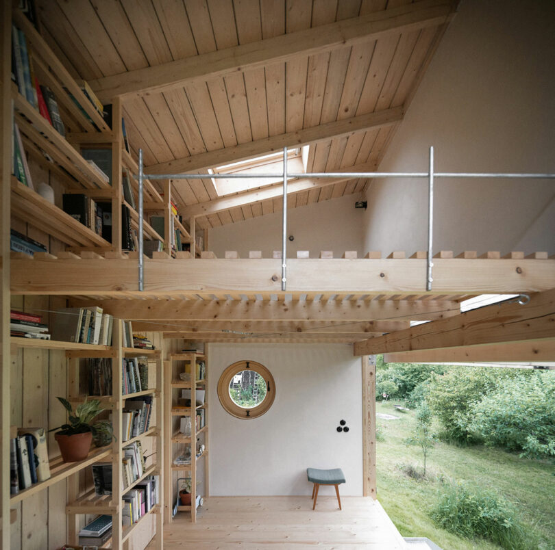 A cozy, wooden, two-level room with bookshelves, a round window, skylights, and an open loft area. A small stool is positioned under the round window, overlooking a green outdoor area.