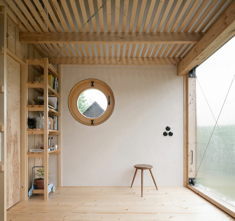 Minimalist wooden room with a circular window, a small shelf with books and plants, and a single wooden stool. The room has large glass-paneled areas and a light wooden ceiling.