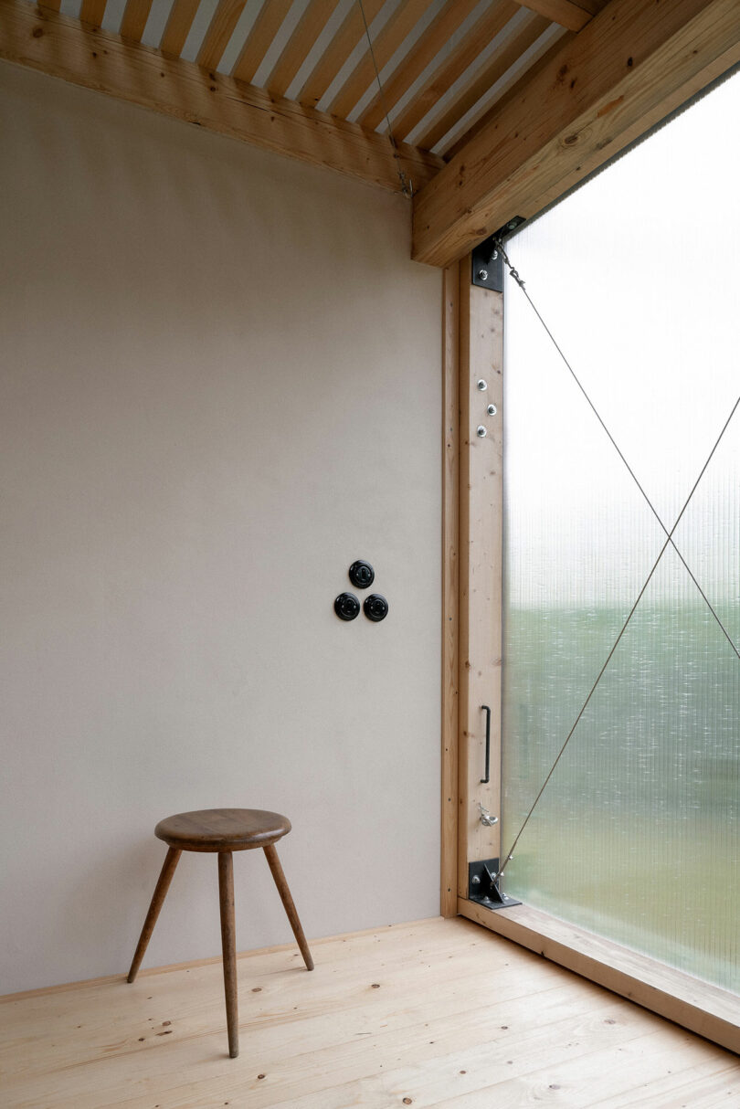 A minimalist room with a wooden stool, three black wall fixtures, a slanted wooden ceiling, and a large transparent window revealing a green outdoor view.