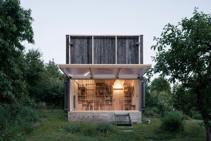 A small modern cabin with a large open doorway showcasing an interior of wooden furniture and bookshelves, situated in a grassy, tree-filled area.