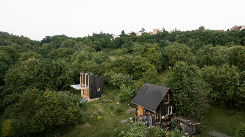 A small, modern house with large windows and dark exterior sits next to a traditional, wooden cabin in a lush green forested area with other buildings visible in the distance.